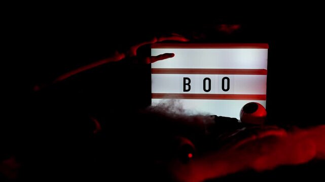 Dark still life featuring illuminated lightbox with BOO message holiday arrangement. Realistic skeleton arm reaches toward display, while decorative spider web creates spooky seasonal atmosphere
