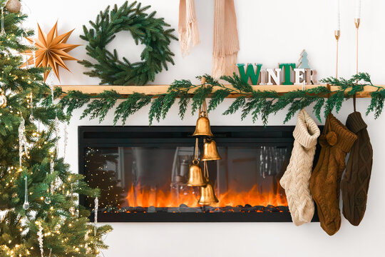Cozy Christmas interior with a decorated fireplace, evergreen garland, wreath, stockings, bells, Christmas tree and stacked firewood. Festive living room, holiday mood, aesthetic decor