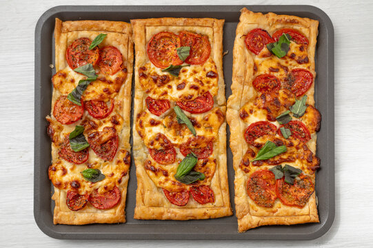 Three rectangular puff pastry, pizza, tomato, cheese, basil, tomato pizza, cheese pizza, savory pas pizzas topped with tomato, melted cheese, basil and herbs, photographed from above on a baking tray.