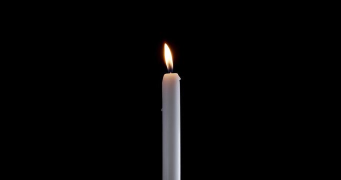 Tall taper candle burning steadily in a dark empty room. A bright flame on this isolated white candle cuts through the shadows. The glowing candle symbolizes peace and spiritual warmth.