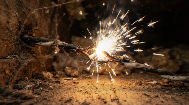 A close-up shot of a damaged electrical cable sparking on a dusty wooden floor, highlighting a dangerous household electrical hazard.