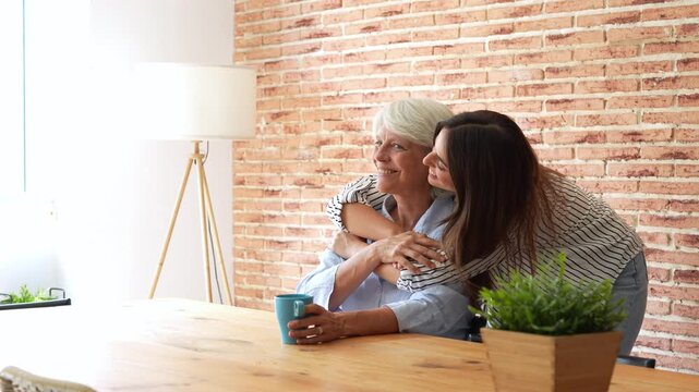 Young adult daughter lovingly surprising and embracing her happy senior mother from behind while she sits at a wooden table, strengthening their beautiful family bond at home together