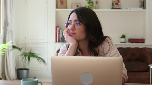 Bored thoughtful young woman with glasses working from home looking out the window, procrastinating on her laptop. A pensive female freelancer feeling unmotivated and distracted