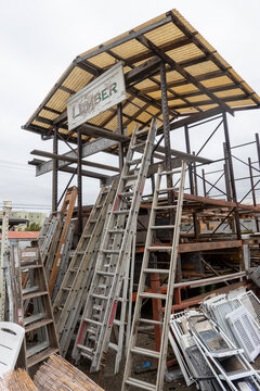 ladders-Multiple assorted ladders lean against a weathered wooden and metal lumber storage structure.