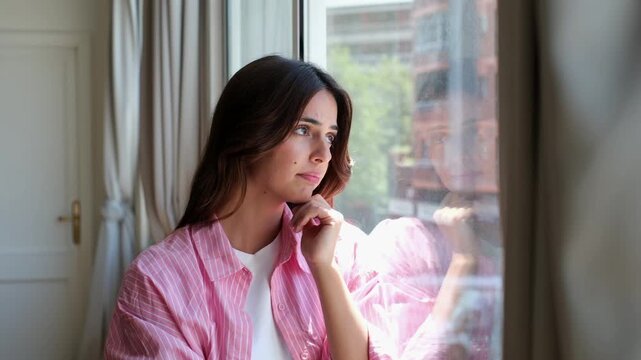 Pensive young woman standing by her apartment window in deep thought, her contemplative gaze fixed on the outside world as she reflects on life, feeling a mix of sadness and hope
