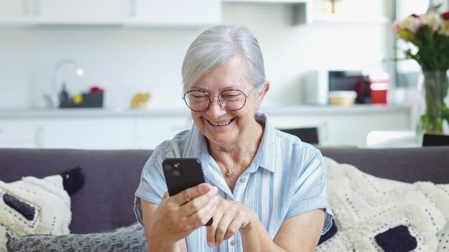 Cheerful senior lady with grey hair and eyeglasses smiling while browsing social media, chatting with family or shopping online using a modern smartphone sitting comfortably on the couch at home	