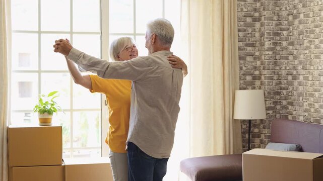 Happy elderly couple celebrating their new apartment by dancing together, surrounded by cardboard moving boxes. Cheerful retired partners enjoy a romantic moment of relocation and new beginnings