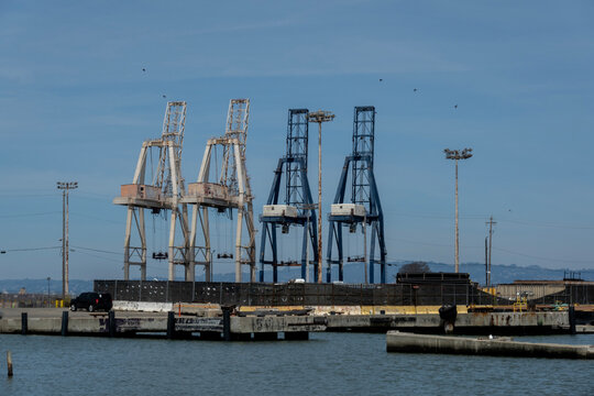 Port Cranes-Large industrial shipping cranes stand tall against a clear blue sky near a concrete harbor pier.