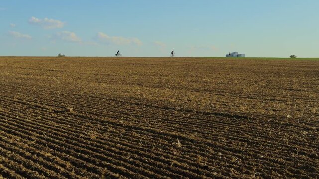 Two cyclists riding along the distant horizon beside a vast cultivated field in Baden-W&uuml;rttemberg, Germany, creating a striking sense of scale and perspective.