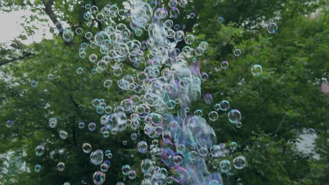 Stream of iridescent soap bubbles floating through the air from a colorful bubble gun against a lush green summer background.