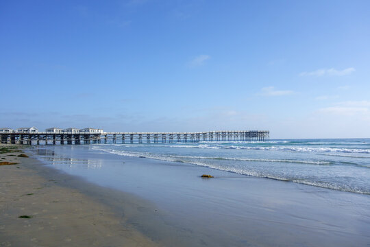 Historic wooden Crystal pier stretching over calm blue ocean under bright summer sky with gentle waves rolling toward sandy shoreline of Pacific beach, San Diego, CA