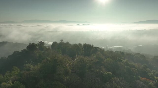 Aerial view of the golden finial of Phra That Chom Kitti Pagoda barely visible above a forested hill, with morning mist and distant mountain silhouettes in Chiang Rai, Thailand, 2023.