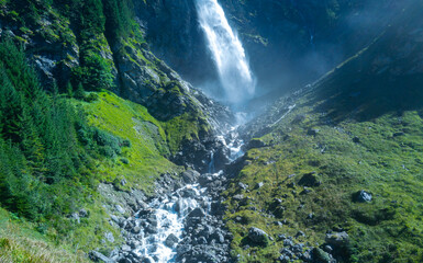 Waterfall feeds a fast mountain stream. Waterfall cascades over rugged mountain rocks. Clear water below waterfall flows downhill. Rocky stream runs below waterfall naturally. © Volodymyr