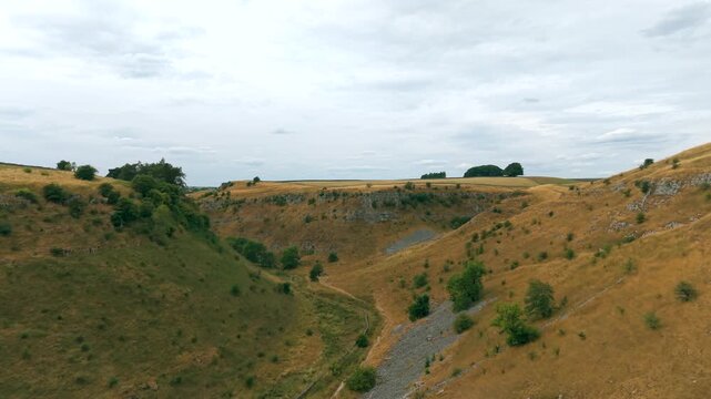 Aerial drone flight descending into a steep, dry limestone valley in the Peak District, featuring dramatic rocky gorge sides, scree slopes, a winding footpath, and rolling hills under an overcast sky