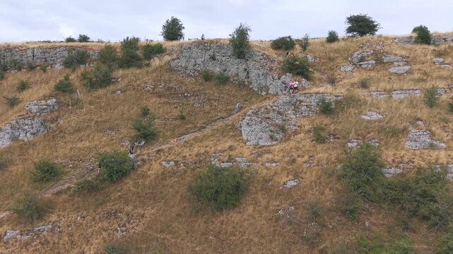 Aerial drone establishing shot of hikers trekking a steep rocky hillside path, revealing a vast scenic rural countryside landscape with green valleys, dry stone walls, and a summer campsite