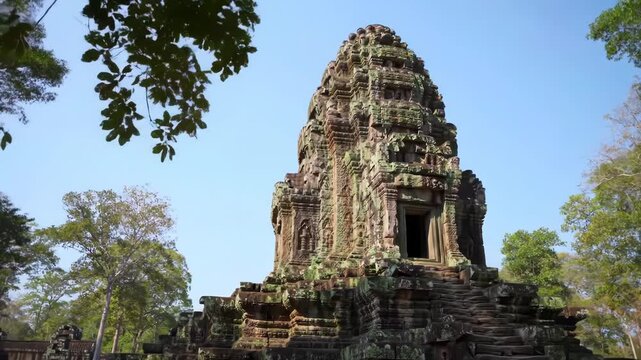 Ancient stone Khmer temple tower at Angkor Archaeological Park in Cambodia featuring intricate sandstone carvings, weathered moss-covered masonry, and tiered architecture under a clear blue sky.
