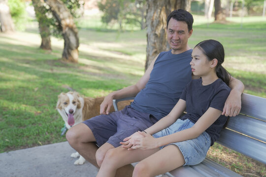 Father daughter dog park family bonding asian man and teenage girl sitting on a bench with their pet dog in a sunny suburban park during a relaxing summer afternoon outdoors.