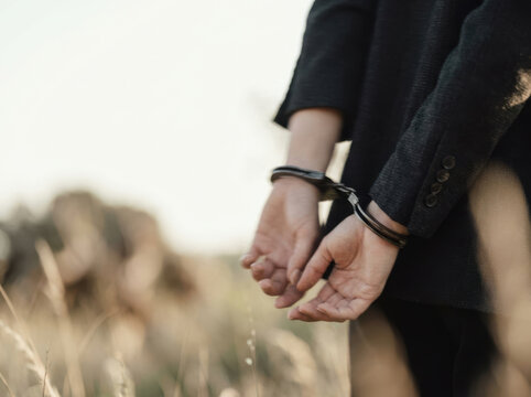 Person with cuffed hands in black jacket standing outdoors in blurred natural background