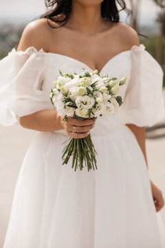 A young woman of Hispanic descent holds a bouquet of white flowers. She wears a white wedding dress with puffed sleeves, standing outdoors with a blurred background.
