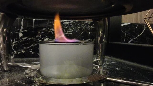 Close‑up of a catering chafing dish fuel burner with visible flame heating a stainless steel serving tray.The setup shows shiny curved metal legs supporting the dish above a cylindrical fuel container