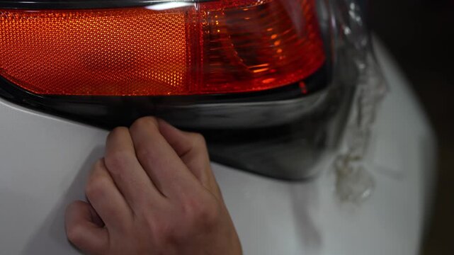 Hand smooths plastic film over car taillight. Fingers press material to ensure full coverage. Clear film adheres tightly to bodywork surface. Professional detail work protects vehicle trim