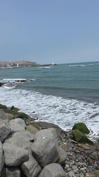 Vertical shot of waves crashing on a rocky shore in Barranco, Lima, Peru. Scenic coastline with a breakwater and lighthouse under a blue sky, background Morro Solar. Natural ocean waves with foam on t