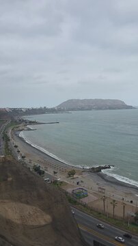 High angle view of the Pacific Ocean coastline in Lima, Peru, featuring the beaches of Barranco and Chorrillos with Morro Solar mountain in the background. Vertical landscape showcasing coastal highwa