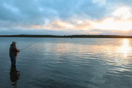An angler stands in shallow water casting a line across the Egegik River at sunrise, surrounded by glowing sky and reflections that fill the calm surface in peaceful Alaskan wilderness.