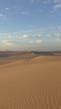 Vertical panning shot from blue sky with scattered clouds over golden sand dunes to the warm setting sun over the Ica desert, Peru. Dynamic landscape transition showing wind patterns, shadows, and ope