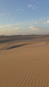 Vertical panning video of golden hour sunset over vast sand dunes in the Ica desert near Huacachina, Peru. Dynamic smooth movement revealing wind-rippled sand textures, shadows, and distant mountain 