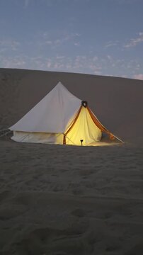 Vertical shot of an illuminated glamping bell tent glowing warmly in the middle of sand dunes at twilight. Scenic luxury desert camping setup under a soft sunset sky, perfect for eco-tourism, travel r