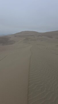 Vertical panning shot of vast sand dunes in the Ica desert near Huacachina, Peru, showing natural wind ripples and vehicle tracks under a moody overcast sky. Ideal for vertical video reels, mobile wal