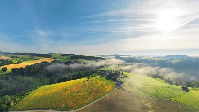 Morning drone footage over the M&uuml;hlviertel in Upper Austria, flying toward the sun above yellow-green fields, forests and a rural village with patches of fog and low clouds.
