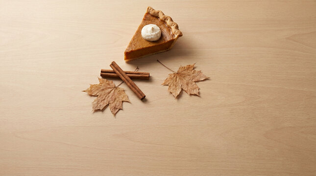 Pumpkin pie slice with cinnamon sticks and maple leaves on a light wood table, a cozy minimalist Thanksgiving top view with copy space for greeting cards