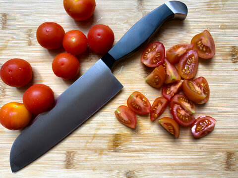 Top view of small cherry tomatoes being sliced with large kitchen knife on bamboo cutting board
