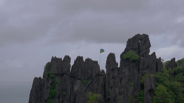 Backward aerial shot revealing jagged tropical rock formations with the Kanaky flag waving in the wind beneath a dark rainy sky in New Caledonia.