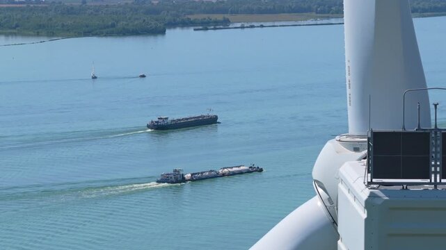 Aerial view from a turning wind turbine showing a blade passing over cargo ships and barges navigating on a wide river in the netherlands