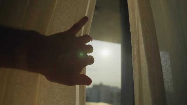 Silhouette of person's hand drawing open window curtain to reveal the sun shining into room in urban city background. Dark room becomes lit by sunlight. Hope and positivity new beginnings and future