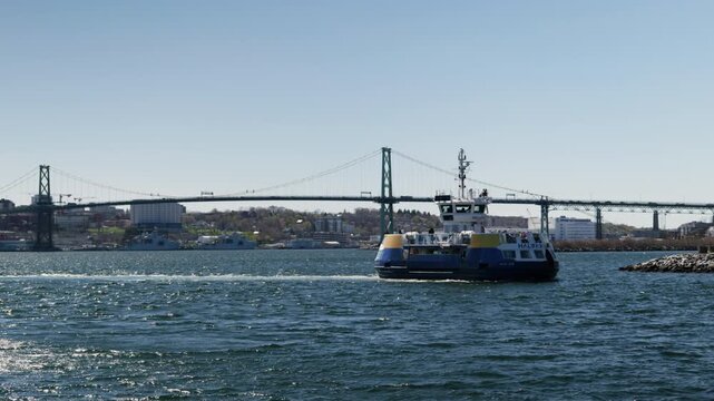 A boat moves through the water near a bridge in the Halifax harbor. The sky is clear, and it is daytime.
