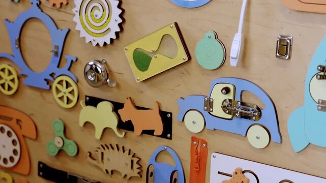 Children explore a wall filled with different objects and shapes to play with. The wall has items like a clock, gears, and animal shapes. Several buttons and latches are included.