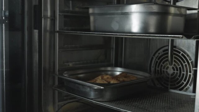 A hand with a blue oven mitt pulls out a tray of baked food from a hot oven. The kitchen shows metal shelves and equipment in the background.