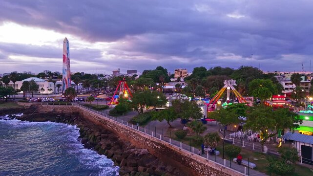 Dusk aerial drone shot flying right over Parque Juan Baron amusement park, Ferris wheel, and coastal avenue with the Obelisco Macho monument in Santo Domingo