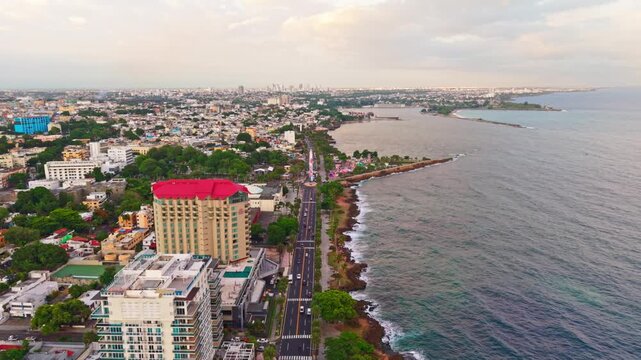 Daytime high-angle aerial drone shot flying forward along coastal highway traffic, rocky coastline, Crowne Plaza hotel, and Obelisco Macho at Santo Domingo Malecon