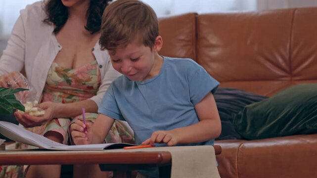 Boy Doing Homework in Notebook Beside Mother on Leather Sofa, Family Learning Activity at Home with Child Student Creativity, Drawing, Concentration, Colored Pencils, Paper, Warm Living Room Interior