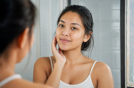 A young woman with a gentle smile examines her reflection in a mirror, her hand lightly touching her cheek, showcasing her natural beauty and the self-care ritual of skincare.