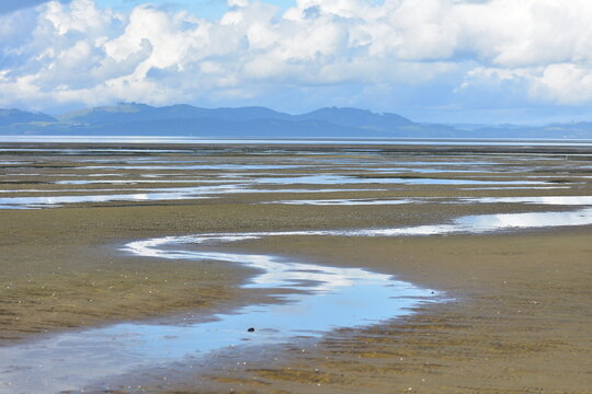 Expansive coastal landscape of tidal mudflats and sand flats exposed at low tide, featuring meandering shallow water channels reflecting a partly...