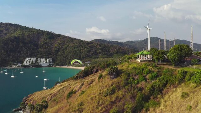 Paraglider soaring above Windmill Viewpoint in Phuket capturing a panoramic scene of Nai Harn Beach, the Andaman Sea with boats, and lush green hills under a bright sky. Active sports and recreation