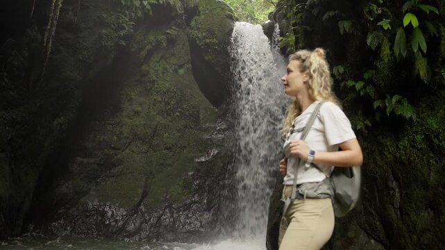 Female explorer with a backpack enjoying the view of a scenic waterfall in a lush tropical jungle