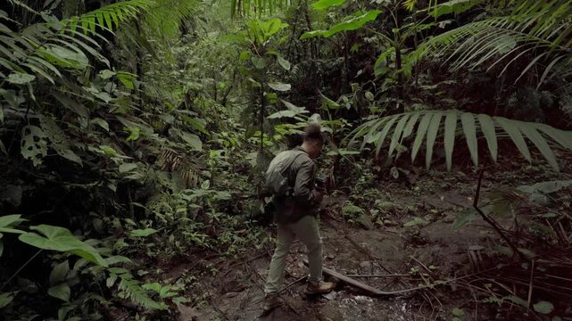 Young man with a backpack and camera exploring a muddy trail in a lush tropical rainforest
