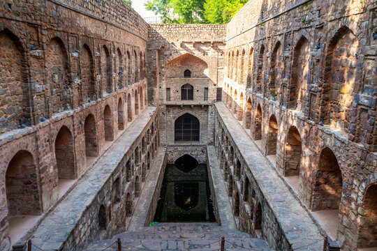 Deep symmetrical view of ancient stone stepwell structure featuring stone arches, reservoir and sunlight cast on historical wall at Agrasen ki Baoli in Delhi, India.
